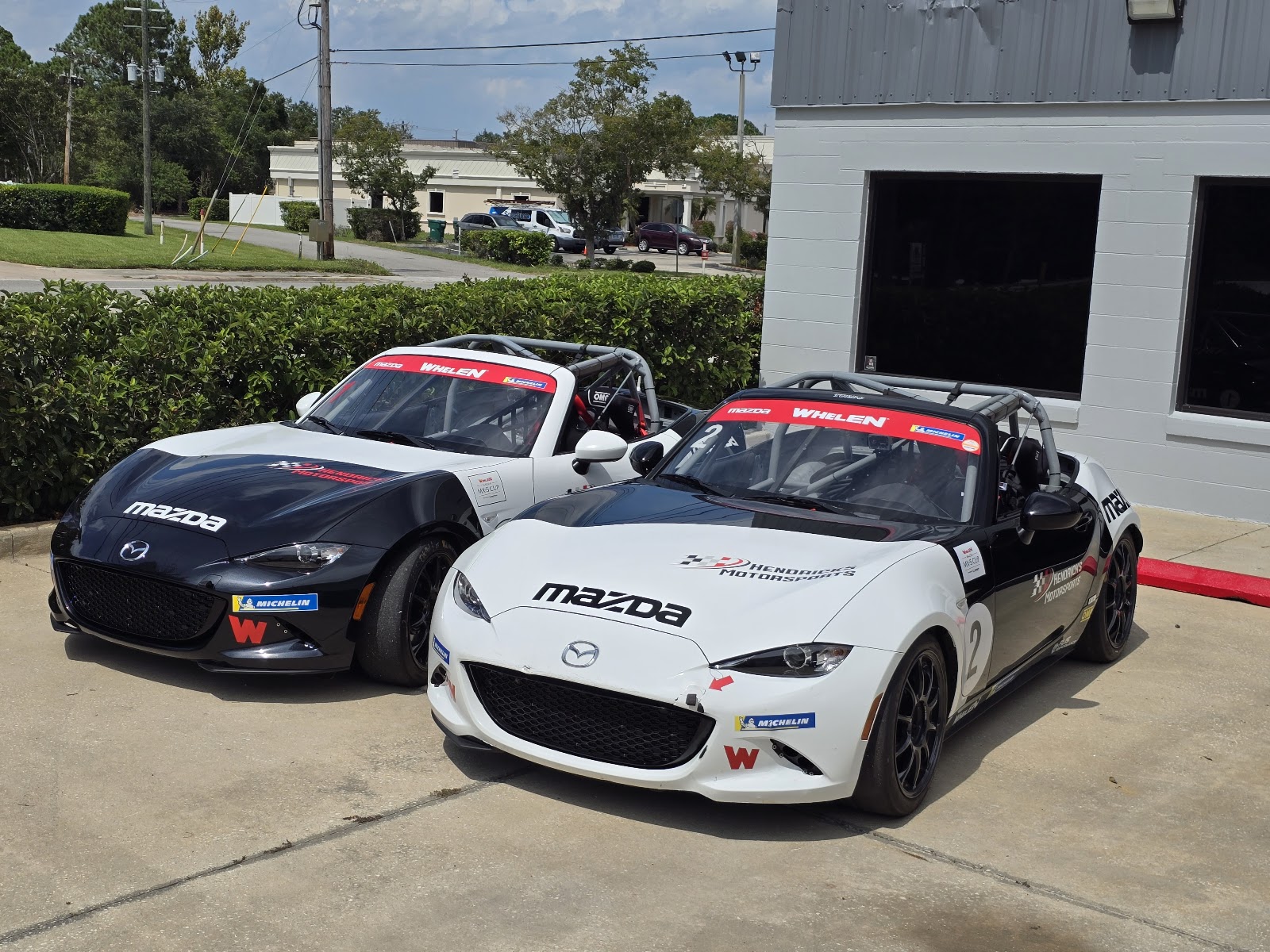Two Hendricks Motorsports Mazda MX-5 Cup race cars (black/white liveries) parked side by side at the Mason Ave shop yard